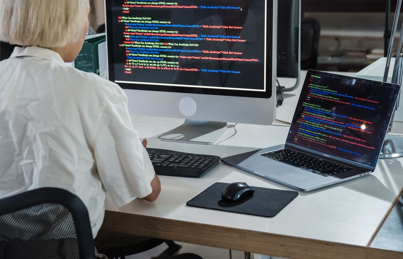 Developer reviewing code on a desktop monitor and laptop in a bright office. Illustrative stock photograph.