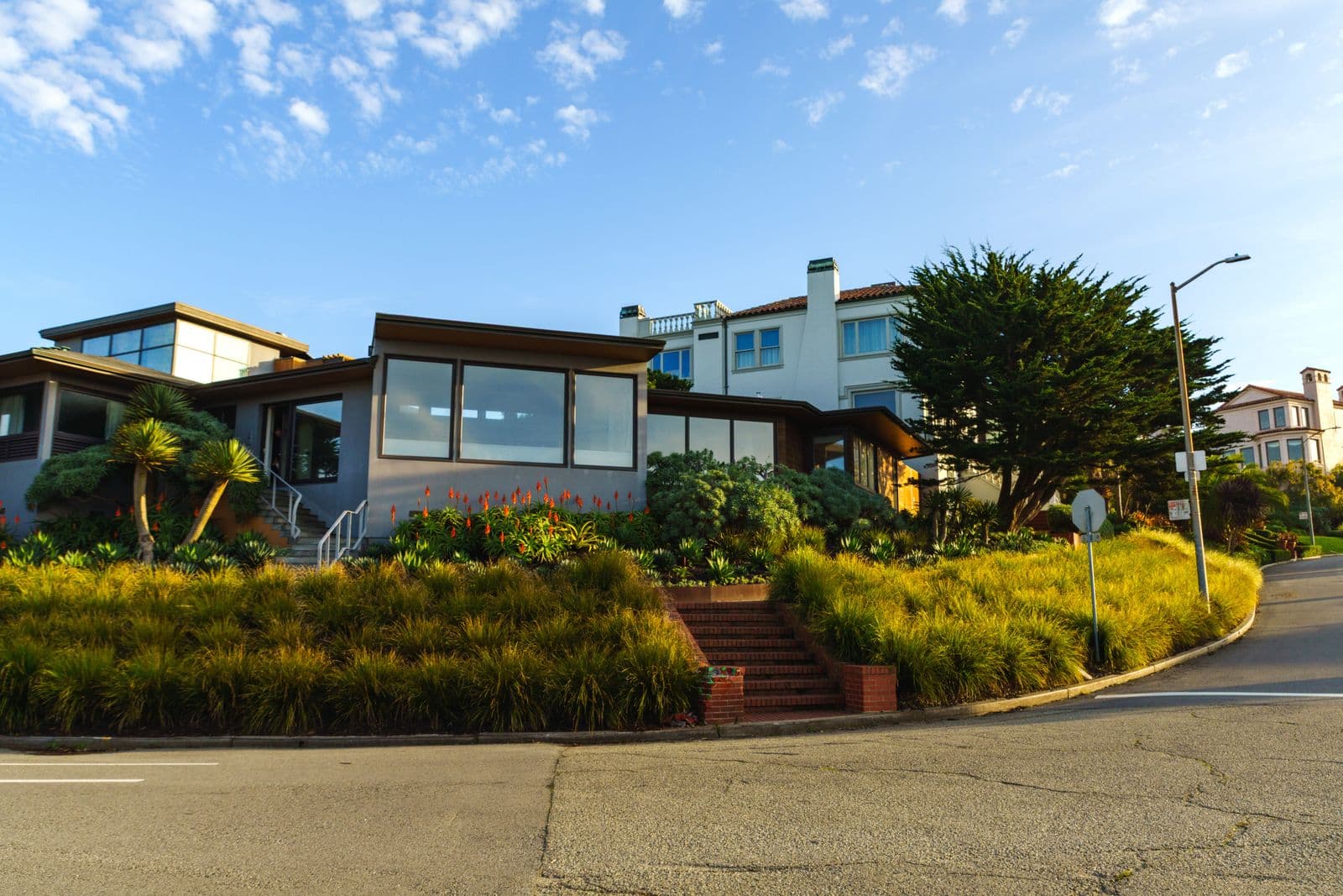 Sunlit coastal home with large windows and lush planting beside a quiet street. Illustrative stock photograph.